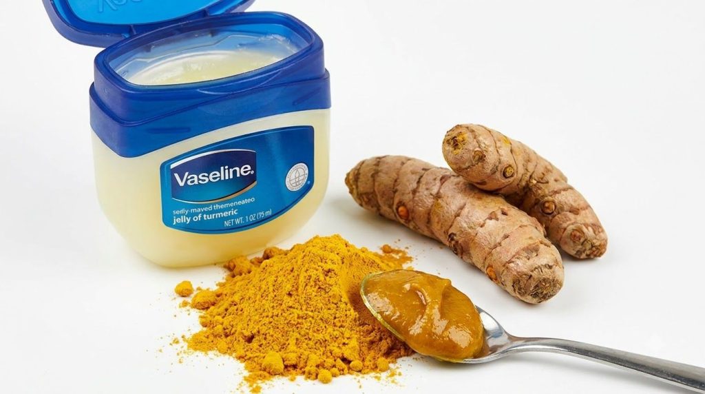 An open jar of Vaseline next to a small pile of turmeric powder and some turmeric roots with a spoon showing a mixture o