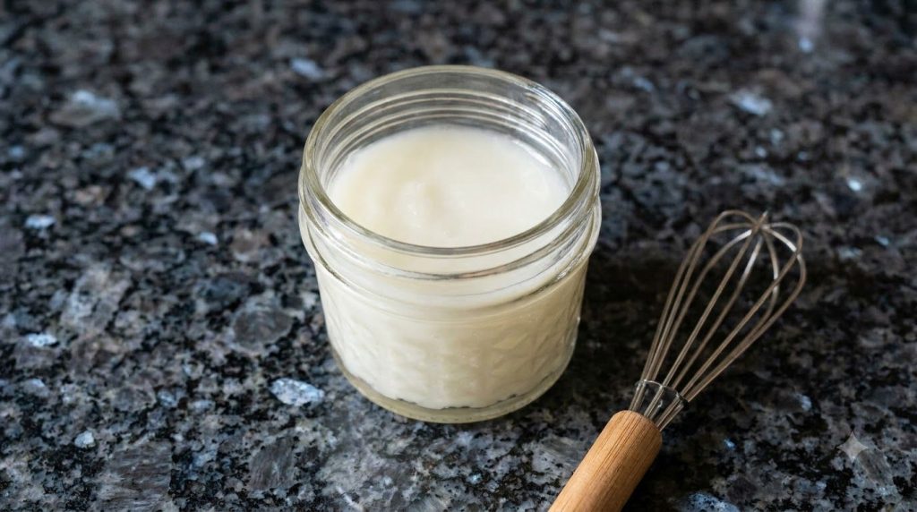A close-up of a glass jar filled with the freshly made creamy white glow gel with a mini whisk resting beside it on a da