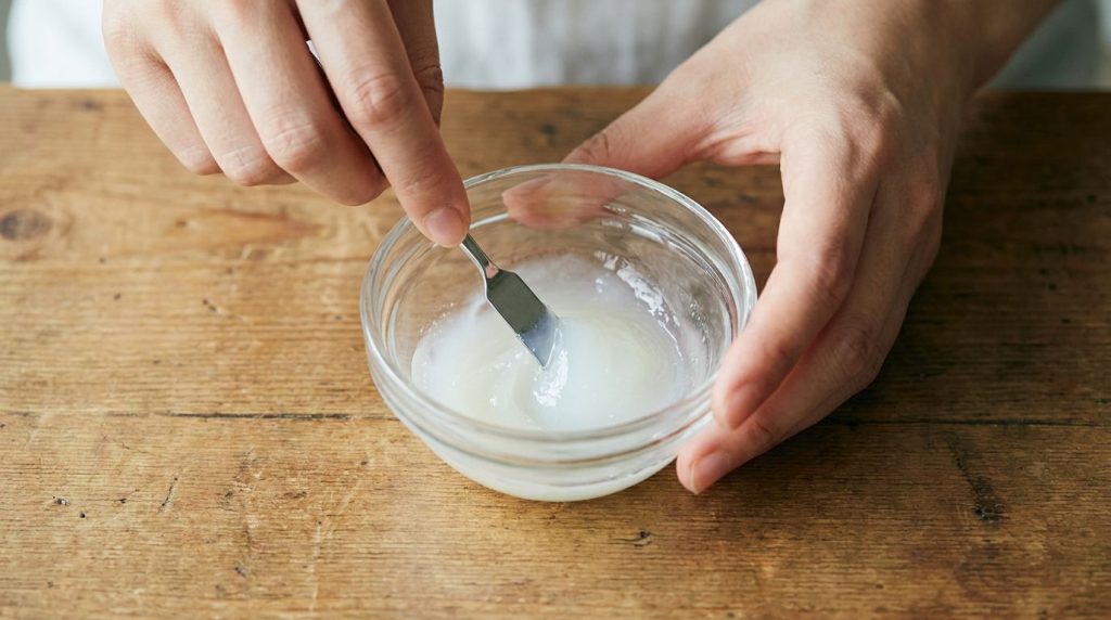 A close-up shot of hands mixing Vaseline and Johnson s Baby Oil in a small bowl with a spoon showing a smooth creamy tex
