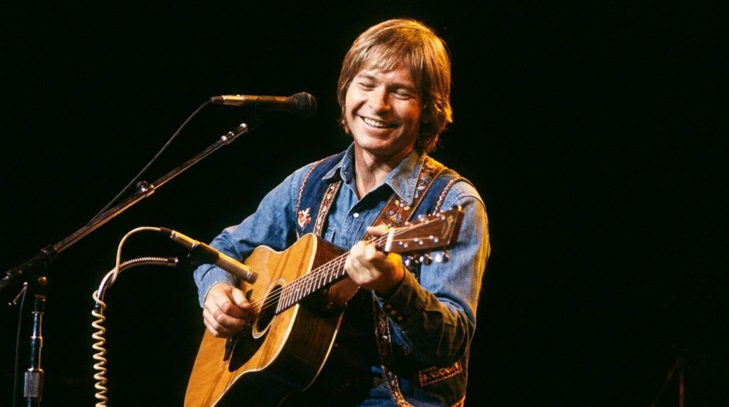 John Denver performing live on stage in the 1970s holding his acoustic guitar with stage lights subtly illuminating him