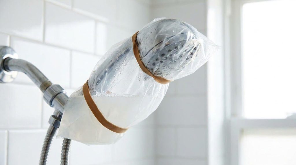 A shower head submerged in a clear plastic bag filled with white distilled vinegar tied securely around the shower arm