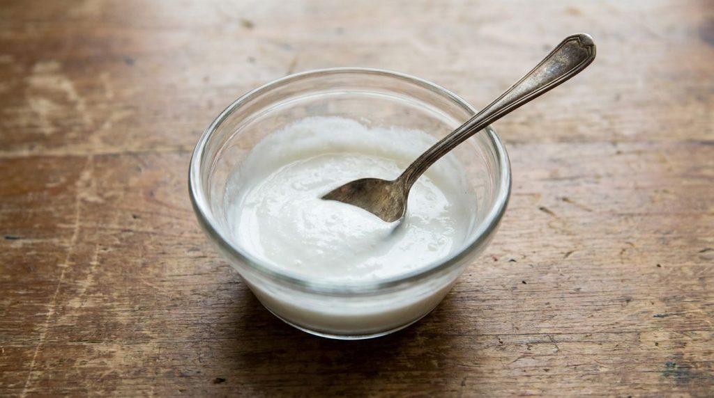 A close-up of a small glass bowl containing a smooth creamy white paste made from crushed aspirin and baby oil with a sp