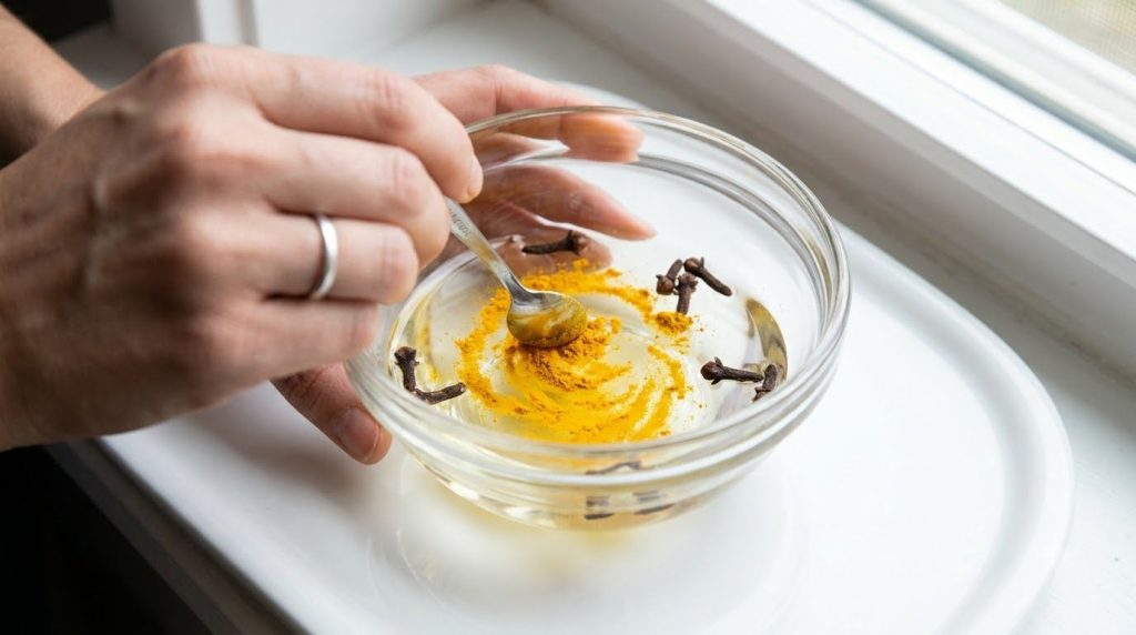 A close-up of a hand mixing turmeric powder into baby oil with cloves in a small glass bowl showing the golden color for