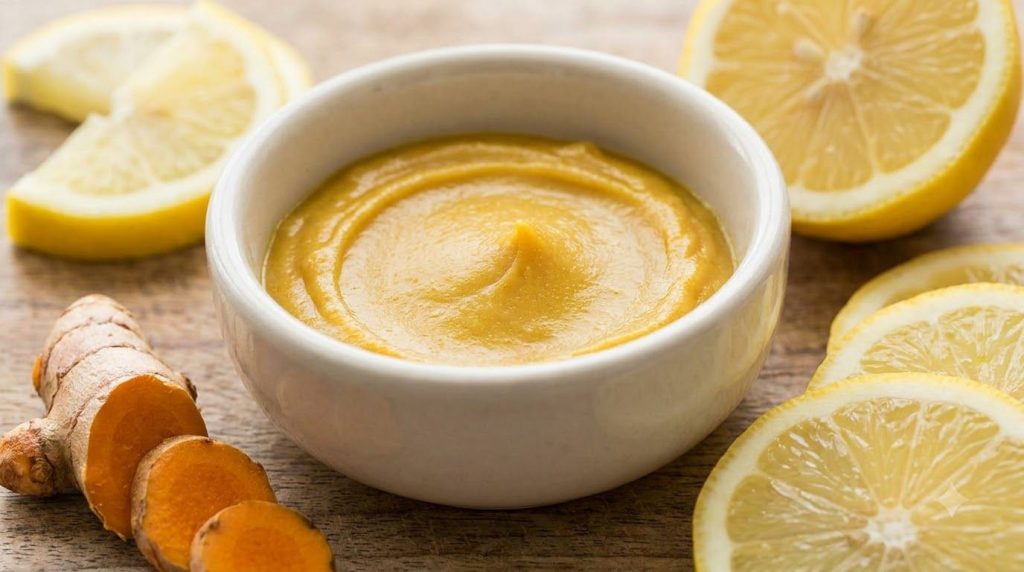 Close-up of a bowl with mixed Vaseline and turmeric paste next to fresh turmeric root and lemon slices