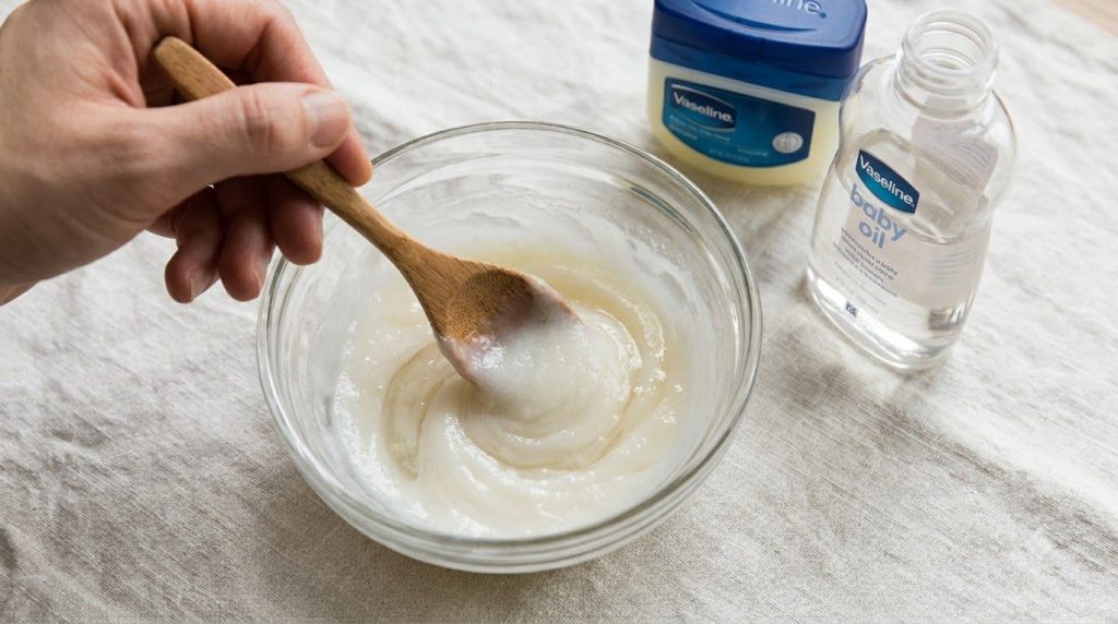 A close-up shot of a small glass bowl with a wooden spoon mixing Vaseline baby oil and baking soda into a smooth creamy 
