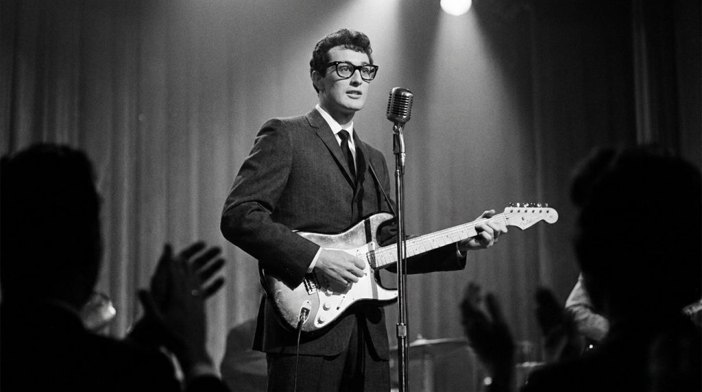 A real archival black and white photo of Buddy Holly wearing his iconic black-rimmed glasses playing a guitar on stage