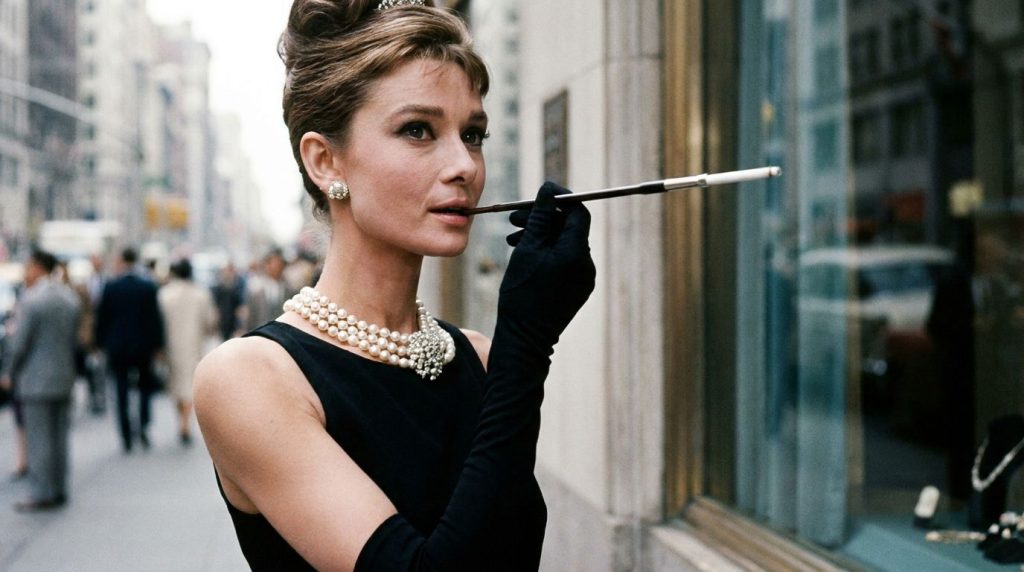 Audrey Hepburn as Holly Golightly in Breakfast at Tiffany s wearing a black dress pearls and holding a cigarette holder 