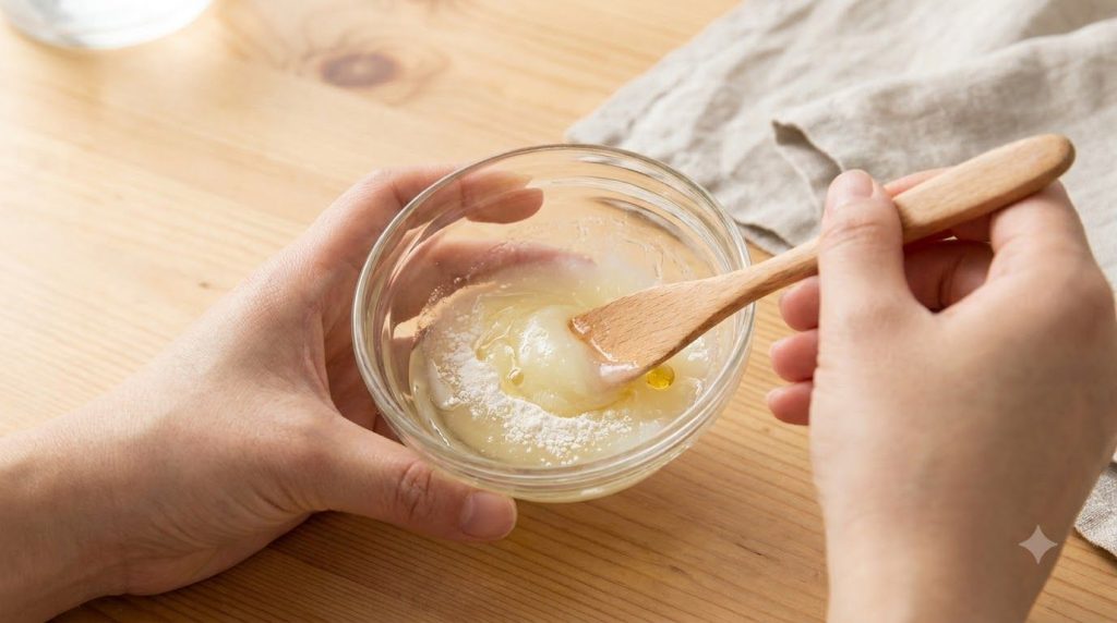 A close-up of hands mixing Vaseline baby oil rice flour and vitamin E oil in a small glass bowl with a mini whisk or spo