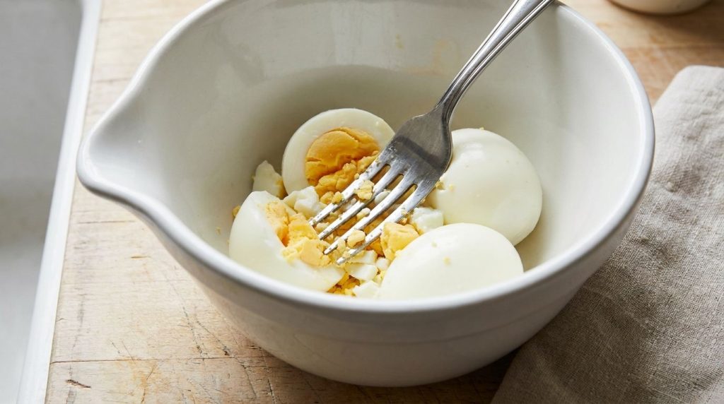 Step-by-step image showing peeled hard-boiled eggs being mashed with a fork in a mixing bowl