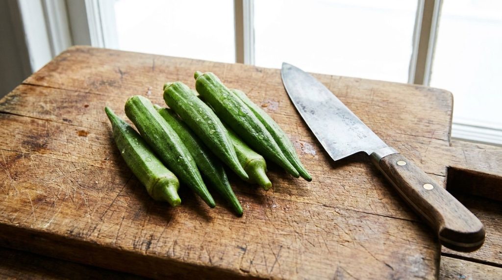 Fresh green okra pods on a cutting board next to a knife ready for preparation