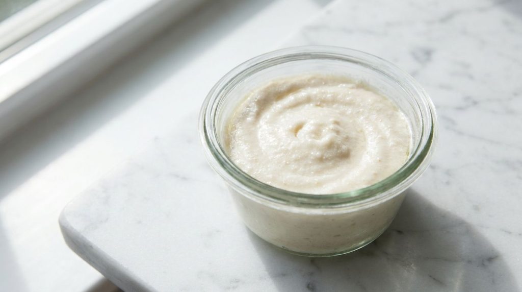A close-up of a small glass bowl on a marble counter containing a creamy white mixture of Vaseline baby oil and baking s