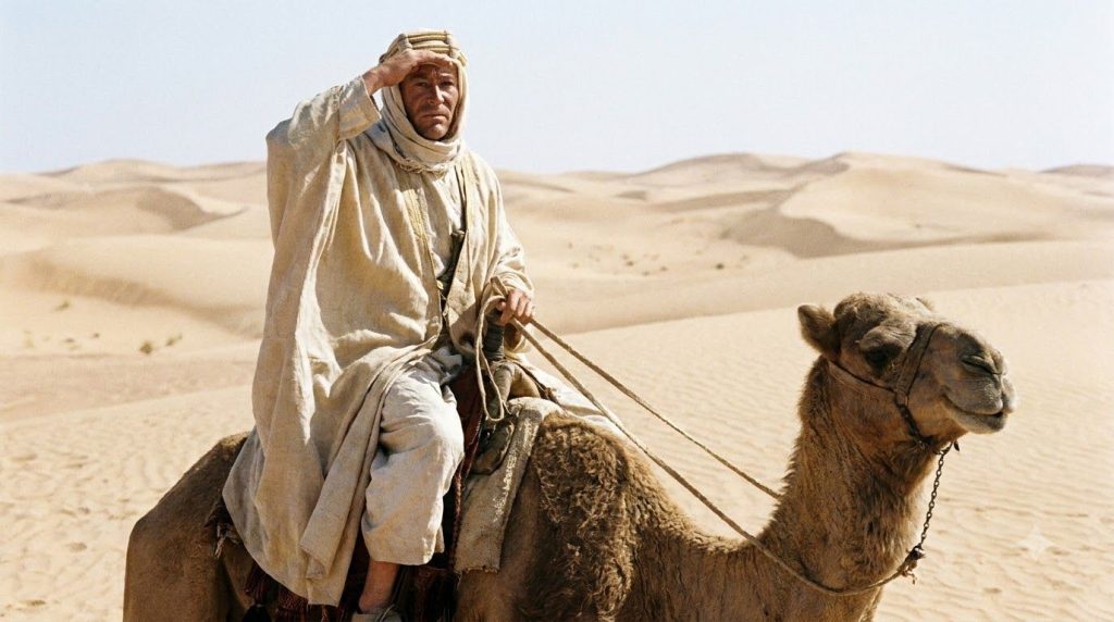 Peter O Toole as T E Lawrence in Lawrence of Arabia wearing desert robes and a determined expression riding a camel acro