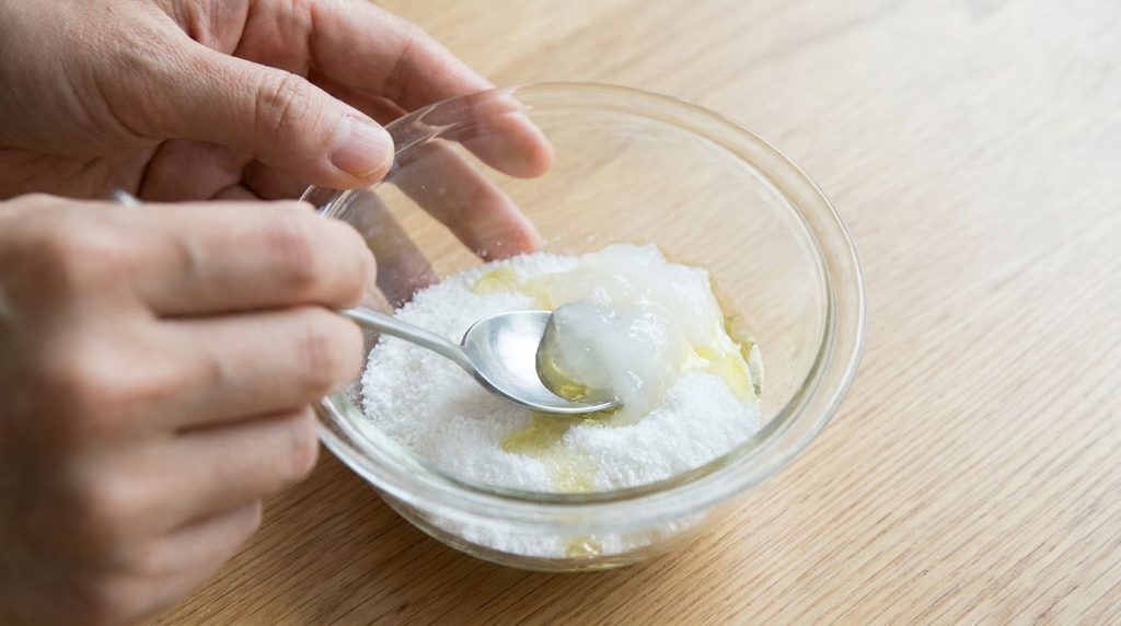 A hand mixing baking soda baby oil and petroleum jelly in a glass bowl to form a creamy paste
