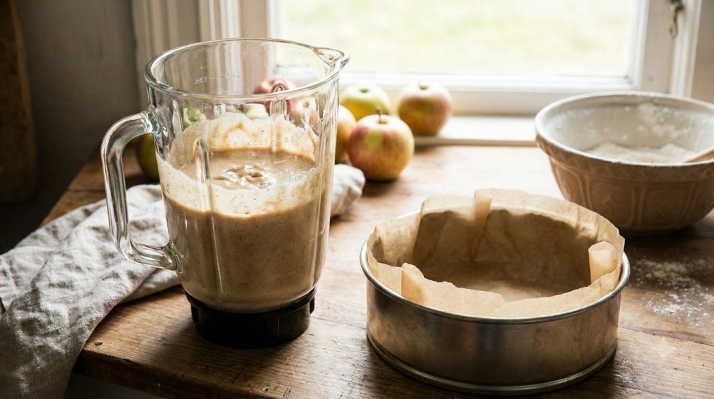 A blender with the apple cake mixture inside next to a prepared baking pan lined with parchment paper on a kitchen count