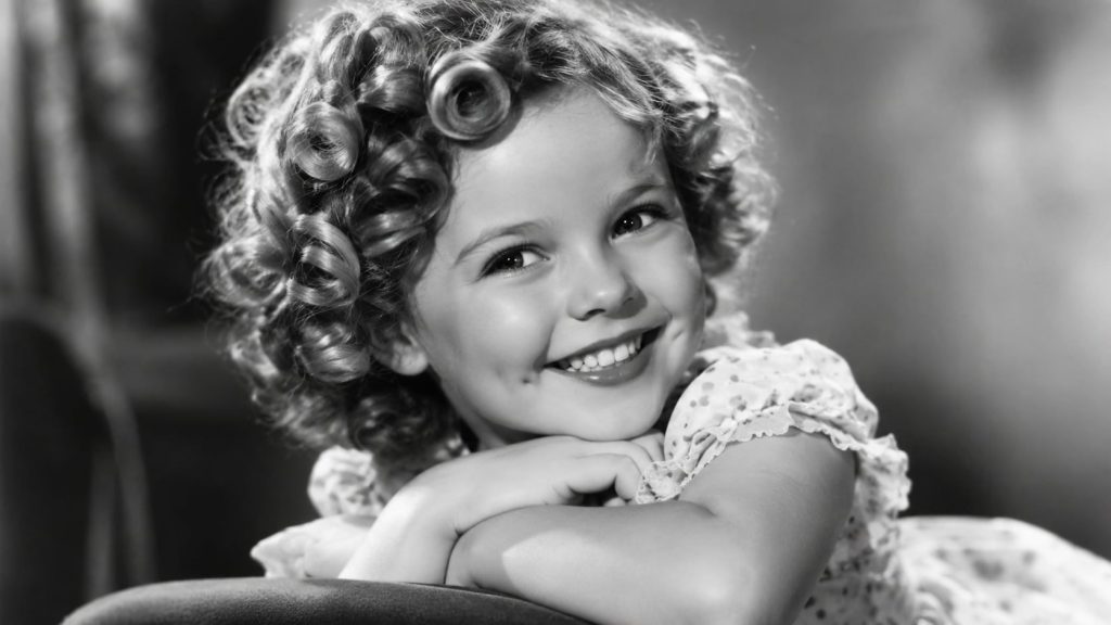 A black and white portrait of a young Shirley Temple smiling widely with her signature curls and dimples