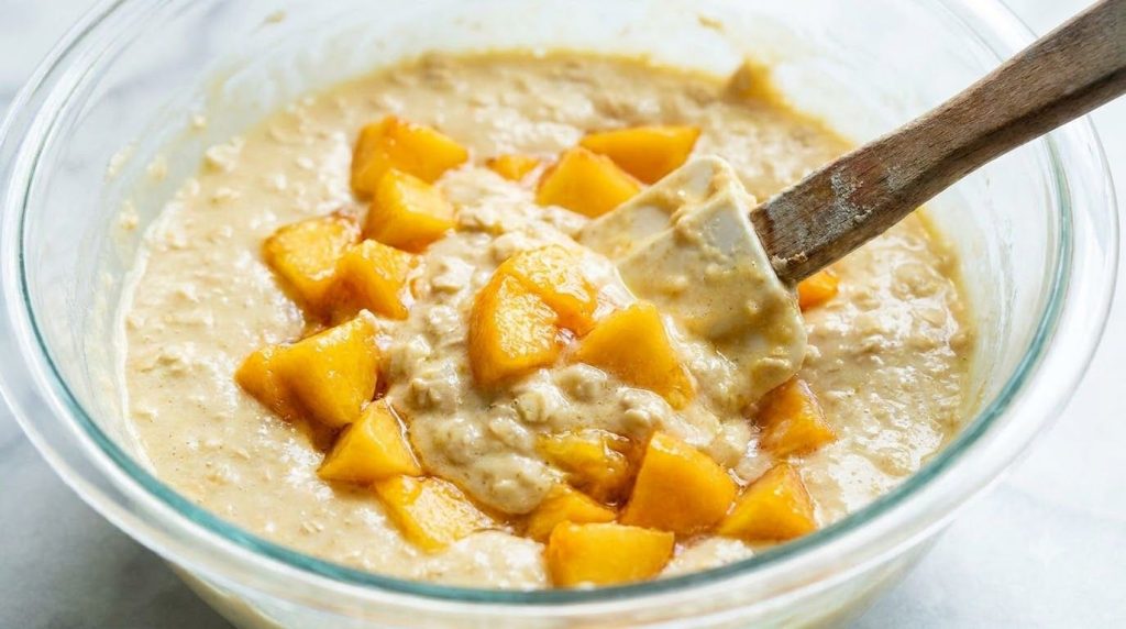 Diced fresh peaches being gently folded into the peach oat cake batter in a mixing bowl showcasing the final stage befor