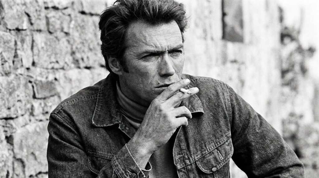 A black and white portrait of a young Clint Eastwood looking stoic and intense perhaps from his Man with No Name era wit