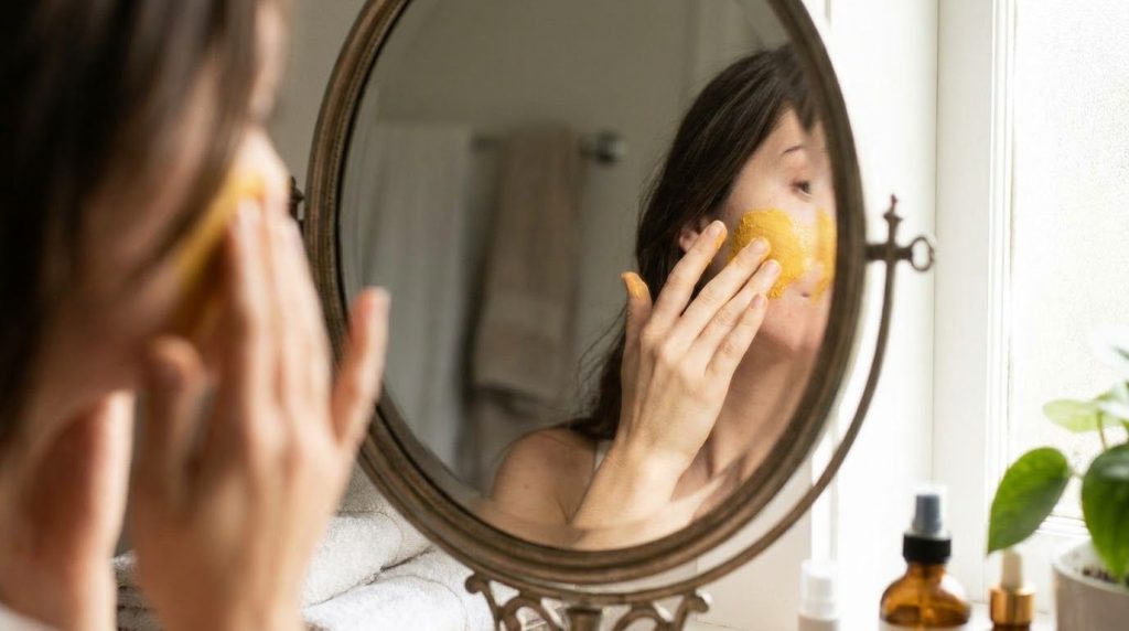 A woman gently applying a yellow turmeric and Vaseline mask to her cheek with her fingertips in a bathroom mirror reflec