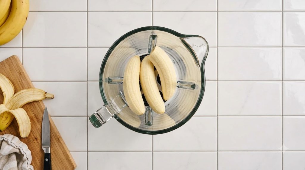 Three peeled bananas in a clear glass blender pitcher on a white tiled surface viewed from above