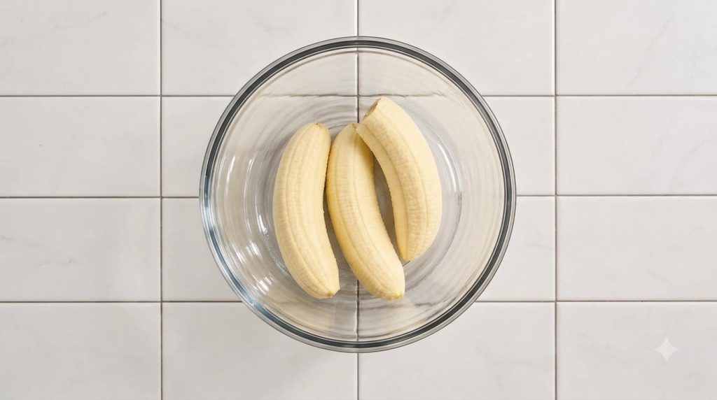 Three peeled ripe bananas in a clear glass mixing bowl on a white tiled surface