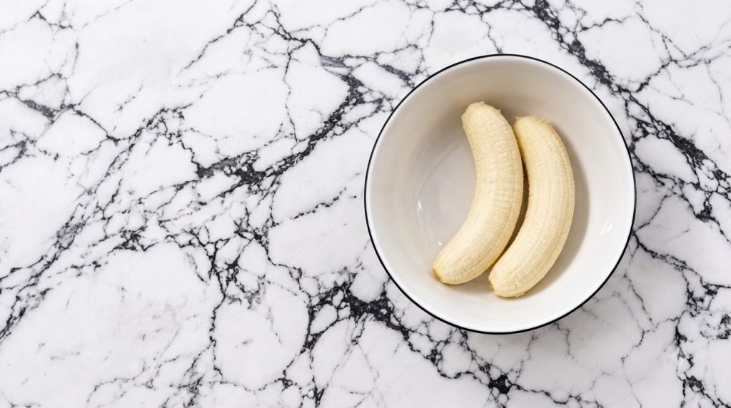 Two peeled bananas resting in a pristine white bowl with a thin black rim centered on a high-contrast white marble backg