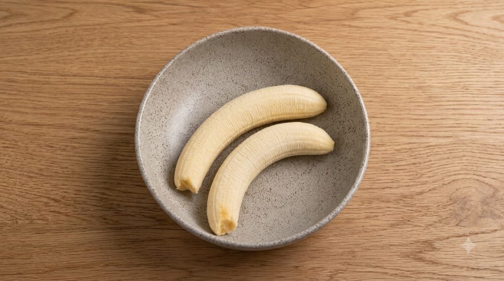 Two perfectly peeled bananas resting in a light gray ceramic bowl on a wooden surface