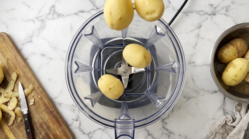 Peeled potatoes being prepared to be blended into a soup with some potatoes suspended above a clear blender pitcher and 