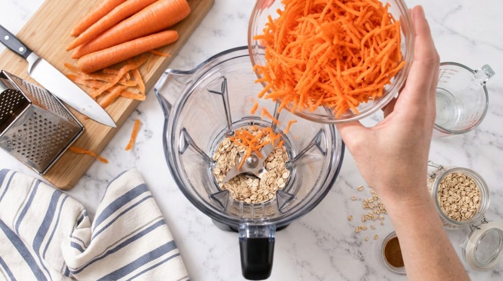 Overhead view of a blender with oats and a hand adding shredded carrots with whole carrots on a cutting board in the bac