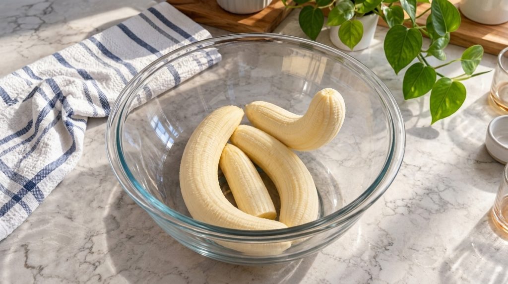 Four peeled ripe bananas in a clear glass mixing bowl on a light marble countertop bathed in bright natural light