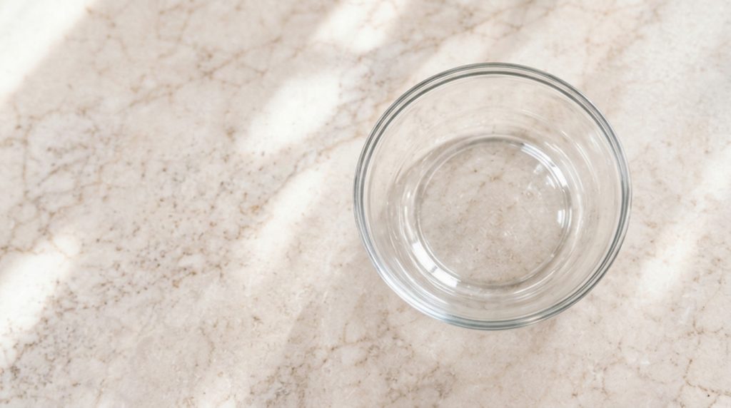 Female hands pouring warm water into a large transparent glass bowl on a wooden kitchen countertop with a dirty greasy c