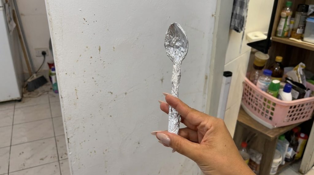 A hand holds a spoon wrapped entirely in crinkled aluminum foil slightly off-center on a light background