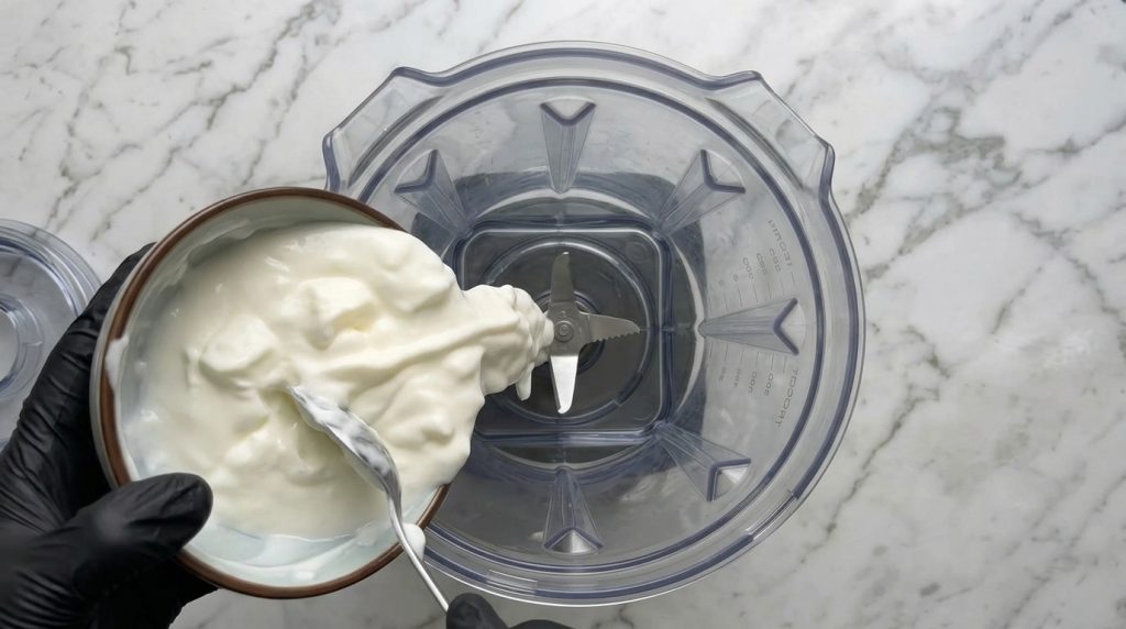 Person holding a bowl of Greek yogurt over a blender ready to add ingredients for a blueberry cake