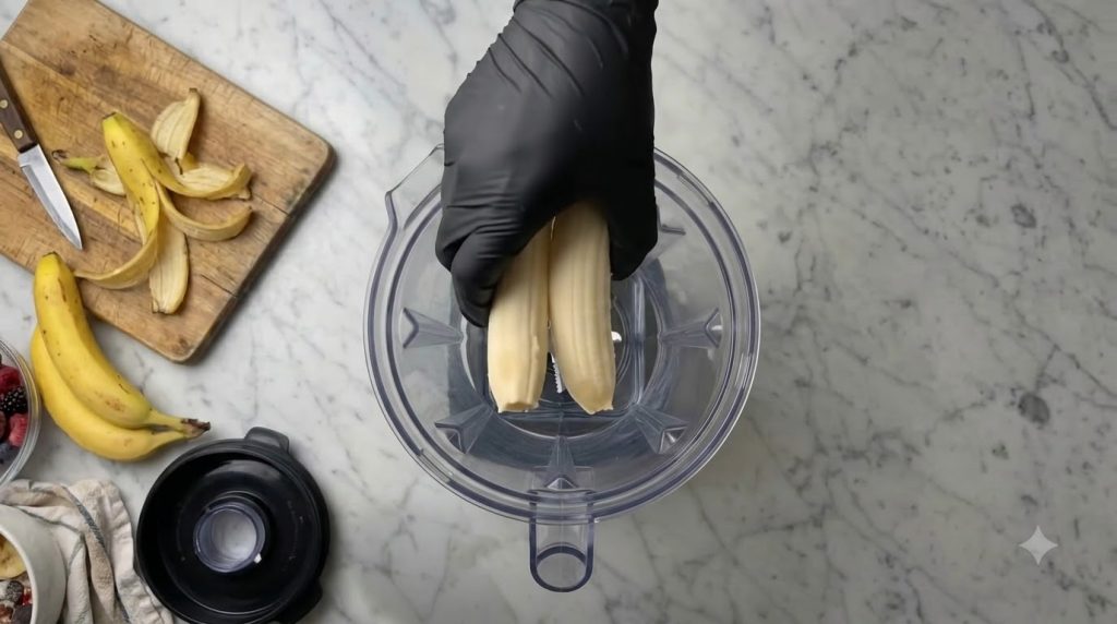 Una mano enguantada de negro sosteniendo dos plátanos pelados sobre una licuadora vacía