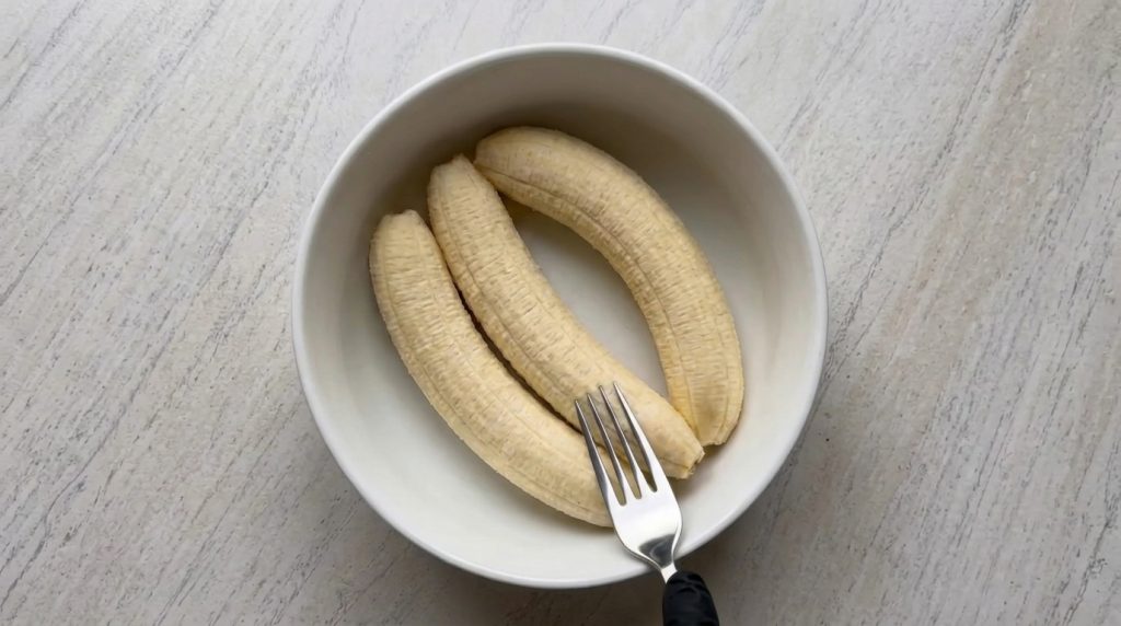 Three peeled ripe bananas in a white ceramic bowl with a hand holding a silver fork gently touching one of the bananas