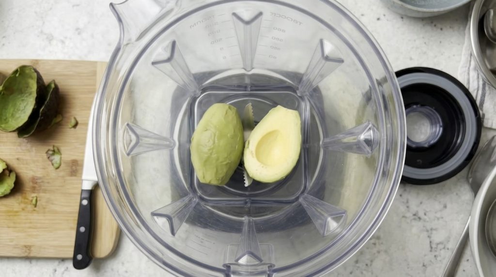 Dos mitades de aguacate fresco una pelada y otra sin hueso dentro de una jarra de licuadora transparente listas para ser