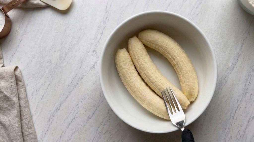 Tres plátanos pelados en un tazón blanco con una mano enguantada sosteniendo un tenedor sobre ellos